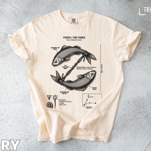 May include: Ivory-colored t-shirt featuring a detailed graphic of two fish intertwined, labeled "Pisces / The Fishes." The design includes astrological symbols and text. The shirt is made of soft material.