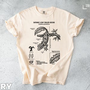 以下が含まれることがあります： アイボリー色のTシャツ。黒と白で描かれた、サタンリーフテイルゲッコーのイラストが特徴で、科学的な注釈が施されています。Tシャツには「SATANIC LEAF-TAILED GECKO」と「Uroplatus phantasticus」の文字が書かれています。