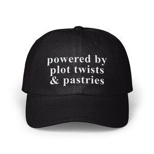 Puede incluir: Gorra de béisbol negra con el texto blanco "powered by plot twists & pastries". La gorra tiene una visera curva y una corona estructurada. Un accesorio casual para los amantes de la lectura y la repostería.