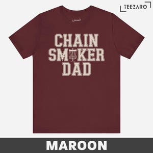 May include: Maroon t-shirt with the text "Chain Smoker Dad" in white with a disc golf disc graphic.