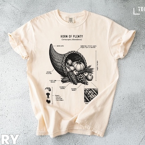 May include: Ivory t-shirt with a detailed illustration of a cornucopia filled with fruits and vegetables. The design includes the text "HORN OF PLENTY" and various labeled elements. The shirt is made of a soft material.
