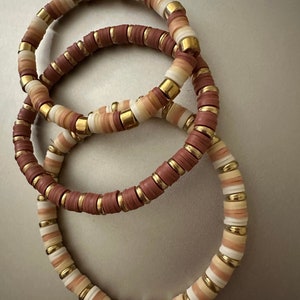 May include: Three stacked bracelets featuring brown, white and gold beads. The bracelets are made from small, round beads with a metallic gold accent.
