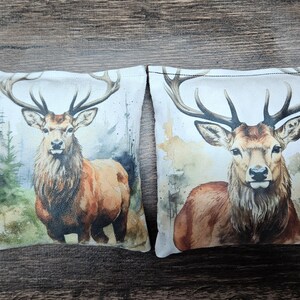 Puede incluir: Dos almohadas de tela blanca con un diseño de acuarela de un ciervo marrón con cuernos en un entorno de bosque.