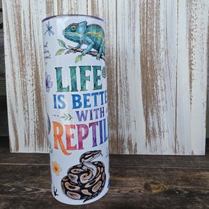 May include: A white cylindrical tumbler featuring a colorful graphic print of a green chameleon on a branch and a coiled brown and black snake. The text reads LIFE IS BETTER WITH REPTILES, accompanied by small illustrations of a dragonfly and flowers.
