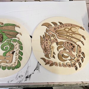 Puede incluir: Dos placas circulares de madera con intrincados diseños de dragones cortados con láser. Un dragón es verde y marrón, el otro es marrón y naranja.