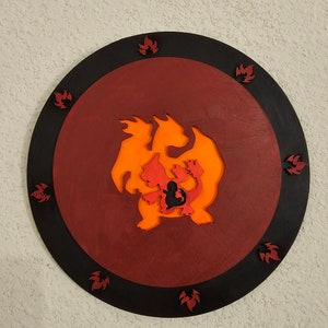 Puede incluir: Una decoración de pared redonda de madera con un anillo exterior negro y un círculo interior rojo. El centro del círculo presenta una silueta de un dragón de tres cabezas que escupe fuego de color naranja y rojo con llamas alrededor de los bordes.