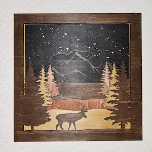 Könnte beinhalten: Wandkunst aus Holz, die eine nächtliche Berglandschaft mit einem Hirsch darstellt. Das Kunstwerk besteht aus geschichtetem Holz mit einem dunklen Nachthimmel, der mit weißen Sternen übersät ist. Die Berge und Bäume sind in Braun- und Beigetönen gehalten, und ein Hirsch ist im Vordergrund silhouettiert.
