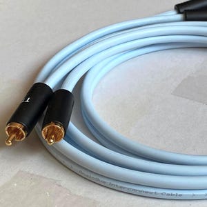 Puede incluir: Cable de interconexión de audio azul claro con conectores negros y puntas doradas. El cable está enrollado y presenta el texto "Analog Interconnect Cable" y "Made in Sweden CE".