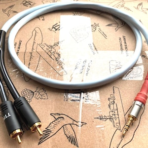 May include: Audio cable with a light gray, rounded cable and black connectors. The cable has a red section near the 3.5mm jack. The connectors are black with gold-colored tips. The cable is coiled on a cardboard surface.