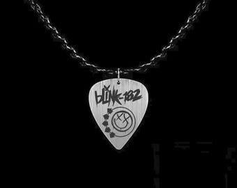 May include: A silver guitar pick pendant with the band name "Blink-182" and a smiley face logo. The pendant hangs from a black chain against a black background. The pick is approximately 1 inch in length.