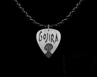 May include: A silver-colored guitar pick pendant with the word "GOJIRA" in a stylized font. Below the text is a silhouette of a head with a tree inside. The pendant hangs from a black chain against a black background.