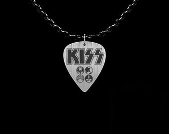 May include: A silver-colored guitar pick pendant with the band name "KISS" in black block letters. The pendant also features four smaller symbols. The pendant hangs from a black chain against a black background.
