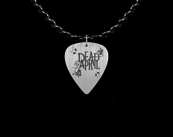 May include: A silver guitar pick pendant with the words "DEAD by APRIL" in black script. The pendant has a textured surface and is adorned with floral designs. It hangs from a dark chain against a black background.