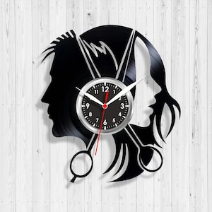 Puede incluir: Un reloj de vinilo negro con siluetas de dos caras y unas tijeras. La esfera del reloj está en el centro, con manecillas blancas y rojas. El fondo es una superficie de madera blanca.