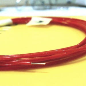 May include: Coiled red wire with a silver tip, resting on a yellow surface. The wire is glossy and appears to be a single strand. A white label is attached to the wire.
