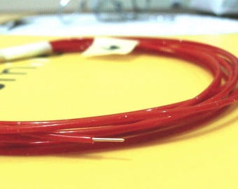 May include: Coiled red wire with a silver tip, resting on a yellow surface. The wire is glossy and appears to be a single strand. A white label is attached to the wire.