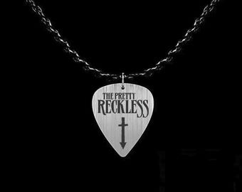 May include: A silver-toned guitar pick pendant hanging from a black chain. The pick features a brushed metal texture with the text THE PRETTY RECKLESS printed in black lettering above a small black downward-pointing cross symbol centered on the surface.