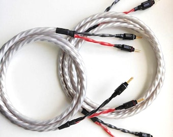 May include: Two coiled speaker cables featuring a braided, translucent silver outer jacket. Each cable splits into two smaller, twisted strands, one black and one red, terminating in gold-plated banana plugs with black plastic housings.