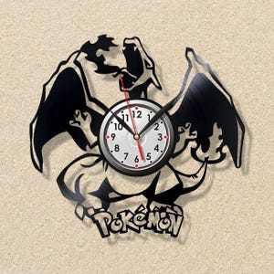 May include: Black vinyl record clock featuring a stylized Charizard design with outstretched wings. The clock face is white with black numbers and hands, and a red second hand. The word "Pokémon" is at the bottom.