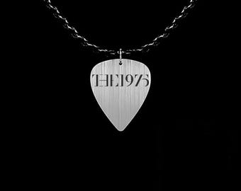 May include: A silver guitar pick pendant with the text "THE 1975" etched on its surface. The pendant hangs from a dark chain against a black background. The pick has a textured, brushed metal appearance.