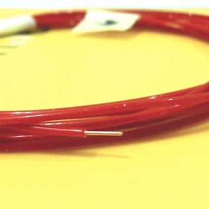 May include: Coiled red wire with a silver tip, on a yellow surface. The wire is a vibrant red colour and appears flexible. A small white label is attached to the coil.