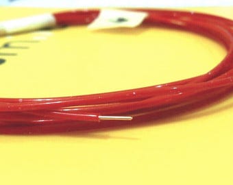 May include: Coiled red wire with a silver tip, resting on a yellow surface. The wire is a vibrant red color and appears to be flexible. A small white label is attached to the coil.
