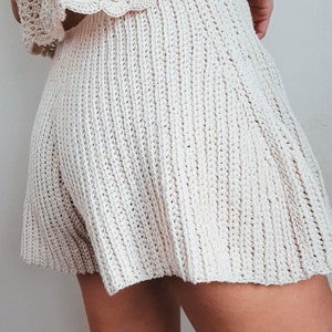 Może przedstawiać: Biała spódnica zrobiona na szydełku z fakturą prążkowaną. Spódnica jest krótka i ma luźny, rozkloszowany kształt.