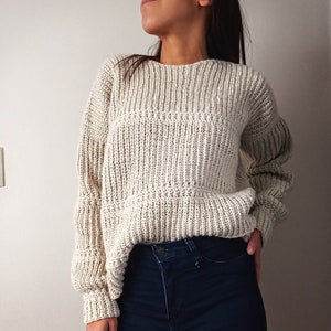 Puede incluir: Un suéter de punto color crema con textura de canalé y un ajuste ligeramente holgado. El suéter tiene un cuello redondo y mangas largas.