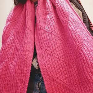 May include: A pink knitted scarf with a diamond pattern. The scarf is folded in half and draped over a person's shoulders.