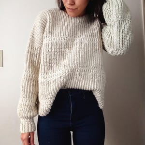 May include: An off-white, long-sleeved, chunky knit sweater featuring a ribbed texture with horizontal bands of decorative stitching across the chest and sleeves, paired with dark blue denim jeans that have a high-waisted fit and visible seams.