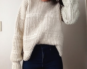May include: An off-white, long-sleeved, chunky knit sweater featuring a ribbed texture with horizontal bands of decorative stitching across the chest and sleeves, paired with dark blue denim jeans that have a high-waisted fit and visible seams.