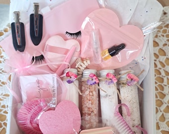 May include: A pink gift box filled with beauty and self-care items. Includes hair clips, heart-shaped items, bath salts in glass tubes, a nail brush, and a card that says "Thank you". The box is lined with tissue paper.