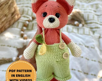 May include: A crocheted fox doll with a peach-colored body, cream-colored accents, and green overalls. The fox has black eyes and a black nose. The overalls have wooden buttons. The text "PDF PATTERN IN ENGLISH WITH VIDEOS" is in a yellow circle.