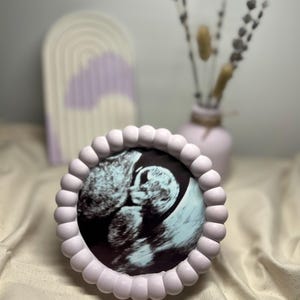 Könnte beinhalten: Ein helllilafarbener Rahmen mit einem perlenbesetzten Rand, der ein Schwarzweiß-Ultraschallbild eines Babys enthält. Der Rahmen steht auf einer weißen Stoffoberfläche.