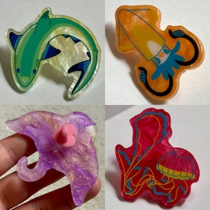 May include: Four colorful acrylic pins featuring sea creatures. The pins include a green shark, an orange squid, a purple dolphin, and a red jellyfish. Each pin has a unique design and vibrant colors, perfect for adding a touch of marine life to any outfit or accessory.