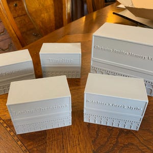 May include: Five light gray rectangular boxes with embossed text: "Old Testament", "Timeline", "Latina Christiana", "D'Aulaire's Greek Myths", and "Christian Studies Memory Verse". The boxes are arranged on a wooden surface.