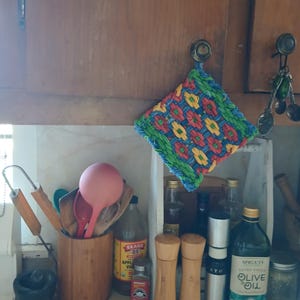 May include: A colorful, woven potholder with a floral pattern hangs from a cabinet knob. Kitchen utensils, including wooden spoons and a pink ladle, sit in a wooden container. Olive oil and vinegar bottles are also visible.