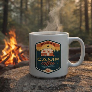 May include: A white ceramic mug with a "Camp Coffee" logo featuring a vintage camper, trees, and the text "Roasted in GA." Steam rises from the coffee. The mug sits on a rock near a campfire and a tent.