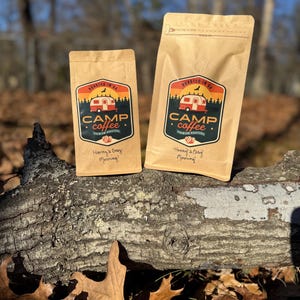 Może przedstawiać: Dwie brązowe papierowe torby Camp Coffee z logo przedstawiającym czerwony kamper, drzewa i napisem "Roasted in GA". Torby mają również napis "Henny's Easy Morning!". Torby leżą na pniu w lesie.