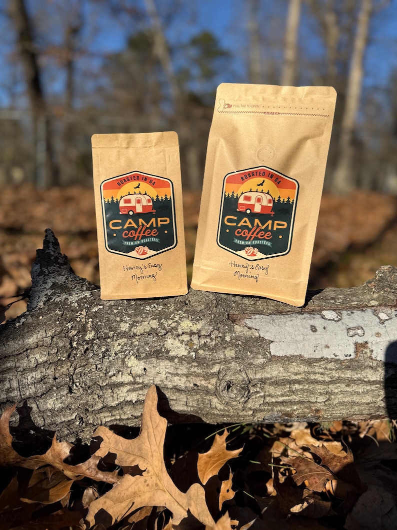May include: Two brown paper bags of Camp Coffee, with a logo featuring a red camper, trees, and the text "Roasted in GA." The bags also have the text "Henny's Easy Morning!" The bags are on a log in a forest setting.
