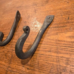 May include: Two hand-forged, dark gray metal hooks. Each hook has a curved design with a rounded end and a flat, rectangular mounting plate with a single hole for screws. The hooks are designed for hanging items.