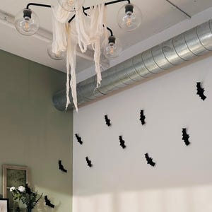 May include: A black metal chandelier with five clear glass globe bulbs, draped with tattered off-white fabric. The wall features multiple small black bat-shaped cutouts arranged in a scattered pattern across the white and sage green surfaces.