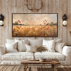 May include: A painting of a field of wildflowers in shades of pink, yellow, and green, with a cloudy sky in the background. The painting is framed in a brown wood frame and is hanging on a wall in a living room setting.