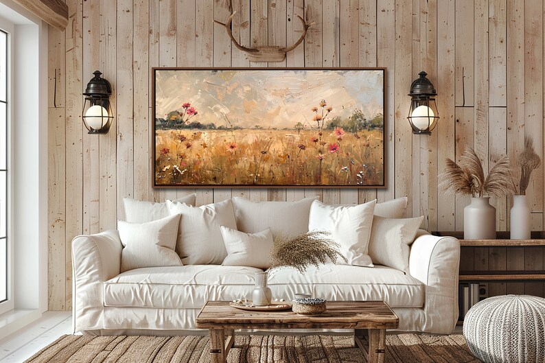 May include: A painting of a field of wildflowers in shades of pink, yellow, and green, with a cloudy sky in the background. The painting is framed in a brown wood frame and is hanging on a wall in a living room setting.