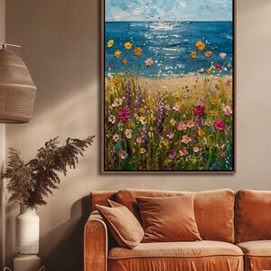 Puede incluir: Pintura al &oacute;leo enmarcada que representa una escena costera con un prado de flores silvestres. El mar y el cielo son azules, con flores amarillas, rosas y moradas. La obra de arte se exhibe sobre un sof&aacute; color &oacute;xido.