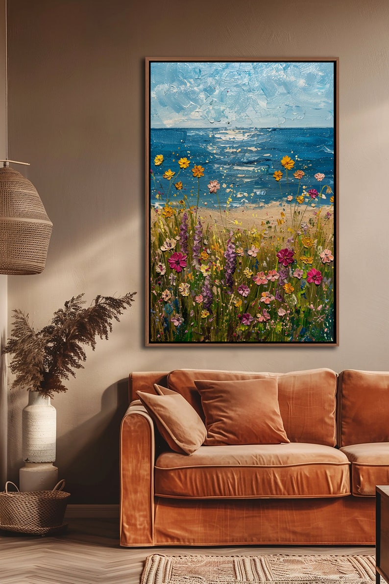 Puede incluir: Pintura al &oacute;leo enmarcada que representa una escena costera con un prado de flores silvestres. El mar y el cielo son azules, con flores amarillas, rosas y moradas. La obra de arte se exhibe sobre un sof&aacute; color &oacute;xido.