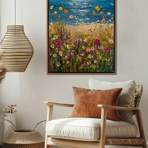 Puede incluir: Pintura al &oacute;leo enmarcada que representa una escena de playa con un mar y cielo azules. Flores silvestres coloridas en tonos rosa, amarillo y morado florecen en primer plano. La obra de arte se exhibe en un sal&oacute;n con una silla y una cesta decorativa.