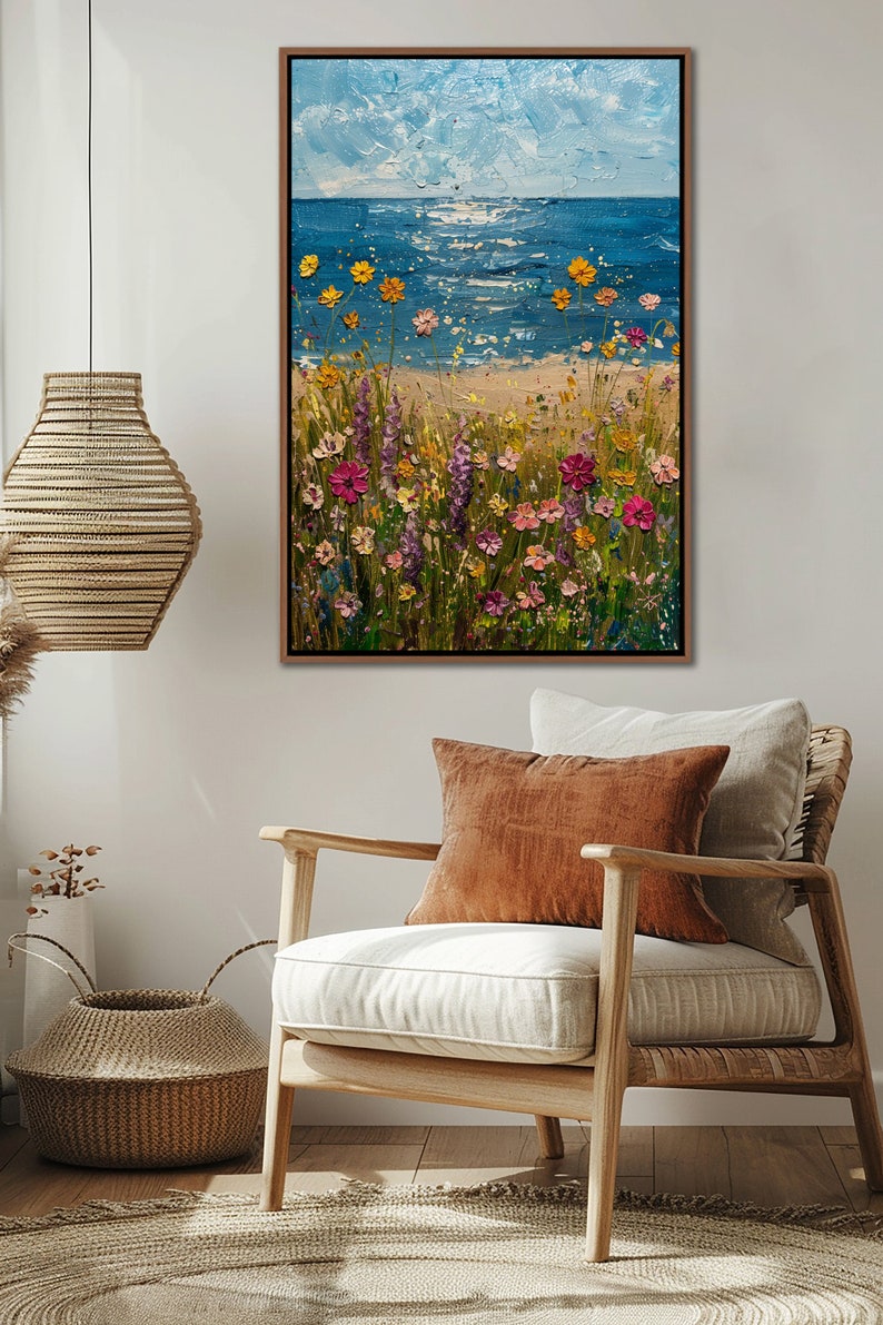 Puede incluir: Pintura al &oacute;leo enmarcada que representa una escena de playa con un mar y cielo azules. Flores silvestres coloridas en tonos rosa, amarillo y morado florecen en primer plano. La obra de arte se exhibe en un sal&oacute;n con una silla y una cesta decorativa.