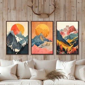May include: Three framed abstract landscape paintings with a mountain range and a large sun in the background. The paintings are in a modern style with a muted color palette of blues, oranges, and reds.