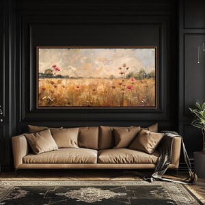 May include: A painting of a field of wildflowers in shades of pink, yellow, and green, with a cloudy sky in the background. The painting is framed and hanging on a black wall in a room with a brown leather sofa and a potted plant.